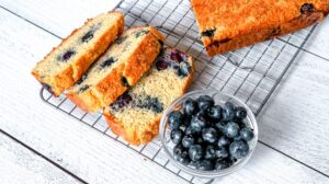 A loaf of Keto Blueberry Bread rests on a cooling rack atop a white wooden surface. Three slices are cut from the loaf, revealing blueberries throughout the bread. A small glass bowl filled with fresh blueberries is placed beside the bread.