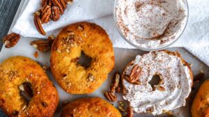 A tabletop displays several freshly baked Keto Pumpkin Pecan Bagels topped with chopped pecans. One bagel is spread with cream cheese and sprinkled with cinnamon. A small bowl of whipped cream, additional pecans, and a white cloth napkin are also placed on the table.