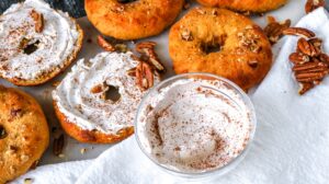 A tabletop displays several freshly baked Keto Pumpkin Pecan Bagels topped with chopped pecans. One bagel is spread with cream cheese and sprinkled with cinnamon. A small bowl of whipped cream, additional pecans, and a white cloth napkin are also placed on the table.