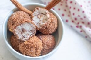 A close-up shot of a bowl of keto churro fat bombs. Some of the bite-sized treats are fully coated in cinnamon sugar, while one is cut open to reveal a creamy cheesecake filling. Two cinnamon sticks are also visible in the bowl.