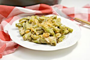 A white plate filled with a creamy Keto Green Bean Casserole topped with pork rind breadcrumbs sits in front of a black skillet with more casserole and a wooden spoon. A red and white checkered cloth and a fork are placed beside the plate.