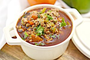 A white ceramic bowl filled with Keto Turkey Chili, featuring ground meat, tomatoes, beans, and garnished with fresh cilantro. The bowl is placed on a wooden surface, with a whole tomato, green bell pepper, and an onion blurred in the background.