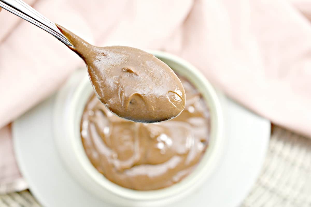 A spoon filled with creamy keto chocolate pudding is held above a bowl of the same pudding. The bowl is placed on a white plate with a light pink cloth in the background. The texture of the pudding appears smooth and rich.