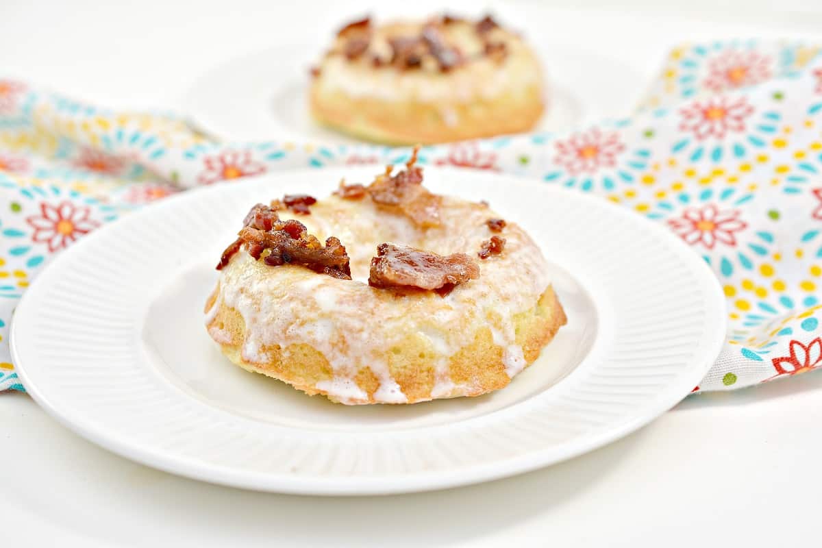 A glazed Keto Maple Donut topped with crispy bacon pieces sits on a white plate. A second donut is partially visible in the background, placed on another white plate. The scene is set on a floral-patterned surface.