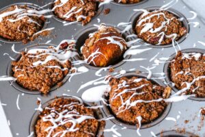 A muffin tin holding several freshly baked Keto Cream Cheese Muffins, each topped with a drizzle of white icing. The muffins have a crumbly, textured topping, and crumbs are scattered in and around the tin. The image is taken from a top-down perspective.