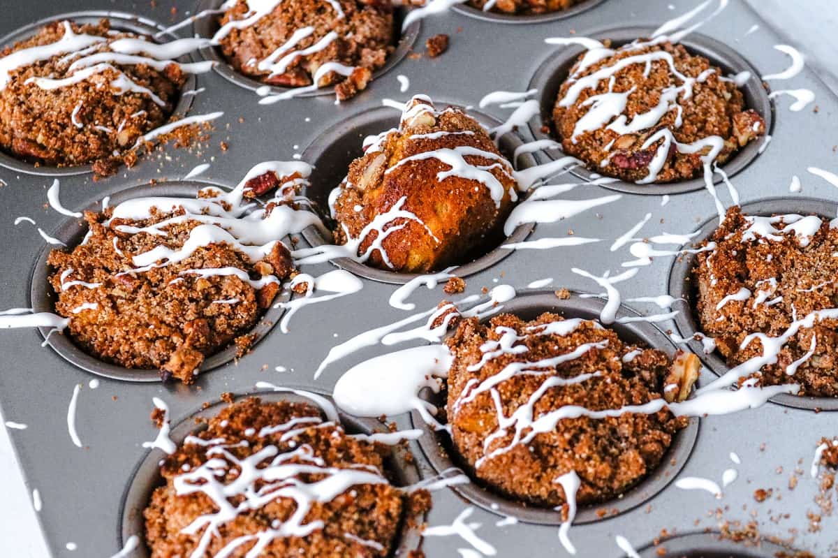 A muffin tin holding several freshly baked Keto Cream Cheese Muffins, each topped with a drizzle of white icing. The muffins have a crumbly, textured topping, and crumbs are scattered in and around the tin. The image is taken from a top-down perspective.