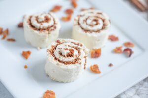 Three Keto Cinnamon Roll Bites are neatly arranged on a square white plate, with small pieces of crispy bacon scattered around them. In the background, there are more cinnamon rolls and a few cinnamon sticks placed on a patterned cloth.