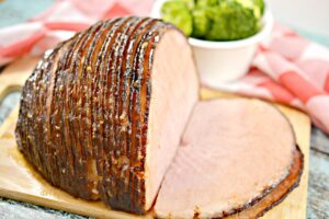 A partially sliced Keto Glazed Ham rests on a wooden cutting board, revealing its pink interior. The ham is neatly scored, with a shiny coating on top. In the background, there's a bowl filled with broccoli florets. A red and white checkered cloth is draped nearby.