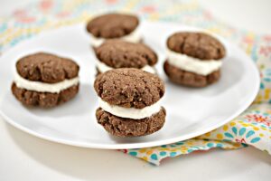 A white plate holds five chocolate Keto Whoopie Pies with cream filling. The plate is placed on a colorful napkin, and the cookies are arranged casually. The focus is on one cookie at the front, clearly showing the filling.