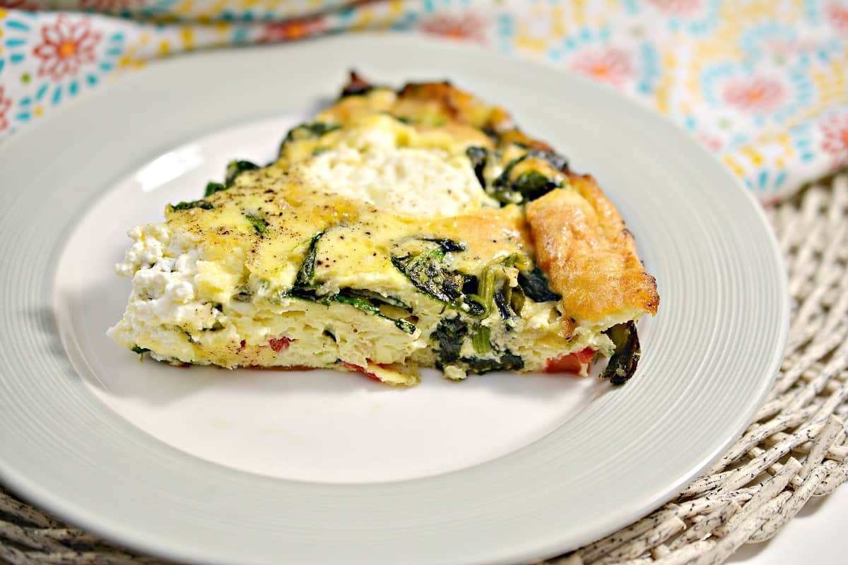 A slice of Roasted Red Pepper and Ricotta Keto Frittata with spinach and ricotta on a white plate. The plate is set on a colorful floral-patterned cloth beside a skillet containing the rest of the frittata. The frittata appears golden brown and garnished with cracked pepper.