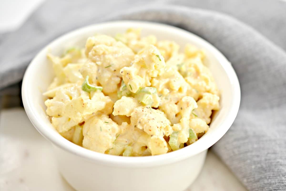 A small white ramekin filled with Cauliflower Potato Salad which consists of diced boiled eggs, chopped celery, and cauliflower florets, garnished with fresh dill. The ingredients are arranged in a colorful and appetizing fashion, ready for mixing or serving.