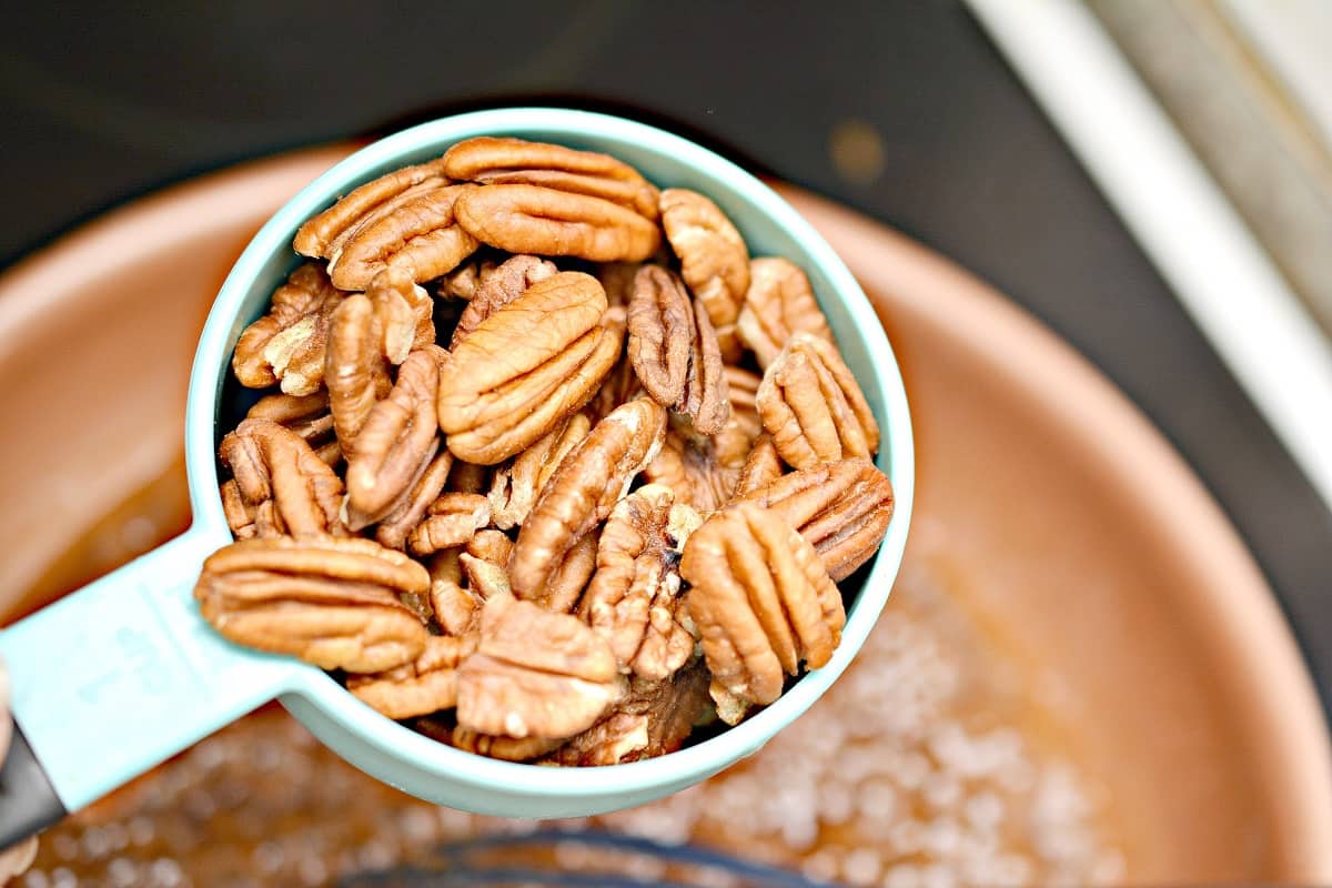 One cup of raw pecans being held over a copper colored frying pan, which contains ingredients for making Keto baked Brie topping.