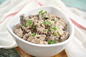 A white bowl filled with Keto Beef Stroganoff, sits on a rustic wooden board. The food appears creamy and savory, garnished with fresh herbs, ready to be served.