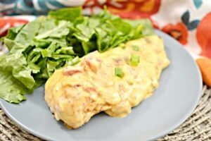 A plate with a serving of green salad and a piece of Queso Chicken covered in a creamy, cheese sauce with pieces of tomatoes and green peppers. The plate is on a woven place mat, and a colorful cloth is partly visible in the background.