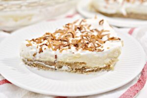 A slice of creamy Keto Coconut Cream Pie topped with toasted coconut flakes sits on a white plate. The pie has multiple layers, including a crust, a creamy middle layer, and a whipped topping. The plate is placed on a white surface with red stripes.