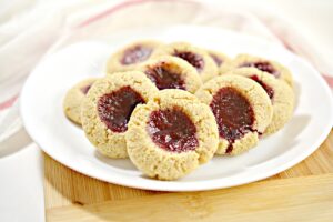 A plate of round, golden-brown Keto Thumbprint Cookies with a dollop of red jam in the center of each. The cookies are arranged in a slightly overlapping manner on a white plate placed on a wooden surface. The background features a blurred striped cloth.
