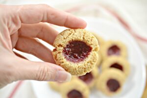 A hand holds a Keto and Gluten Free Thumbprint Cookie, in the background there is a white plate with additional cookies. And a white cloth napkin that has a pink stripe.