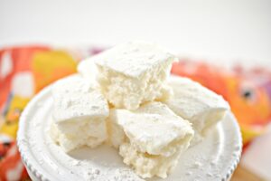 A white plate with a pedestal base holds a stack of white, Keto Marshmallows. A colorful, blurred napkin can be seen behind the plate.