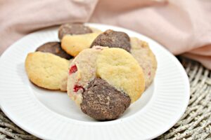 A plate with four Keto Neapolitan Cookies, each divided into three sections: one vanilla, one chocolate, and one with strawberry. The cookies are placed on a white ribbed plate, which sits on a wicker surface with a soft pink cloth in the background.