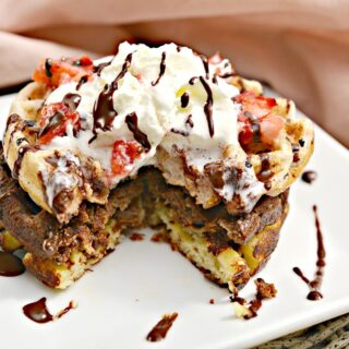 Keto Neapolitan Chaffles topped with whipped cream, strawberry pieces, and drizzled with chocolate syrup. The stack consists of both chocolate, strawberry and plain waffles, presented on a white square plate with a pink cloth in the background. There is a triangular piece that has been cut from the center of the waffles to reveal the tender interior.