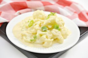 A white plate filled with creamy Keto Cauliflower Mac and Cheese, garnished with chopped green onions. The plate is resting on a black square tile, with a red and white checkered cloth in the background.