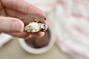 A half-eaten Copycat Keto Almond Joy Bite revealing its fluffy coconut white center, held between fingers over a soft pink-striped background.