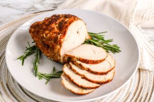 An Air Fryer Turkey Breast is partially sliced on a round white plate. The meat appears juicy and is garnished with sprigs of fresh rosemary. The plate lies on a woven placemat with a beige cloth napkin beside it. The background is a marble surface.