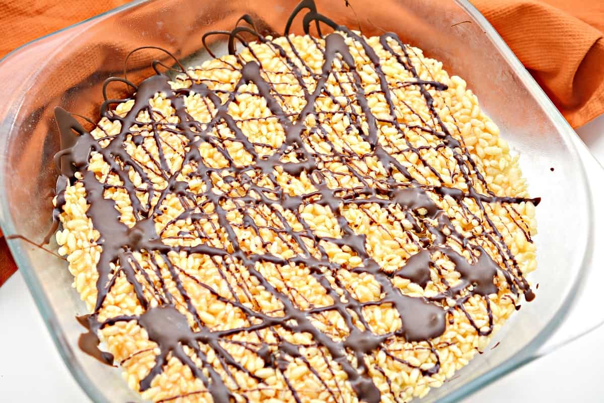 Keto Halloween Rice Krispie Treats pressed into a square glass bowl and decorated with chocolate zigzags, which is situated on a white countertop. There is an orange cloth napkin at the top of the frame.