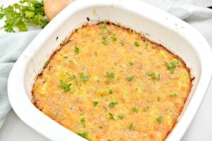 A baked Keto Squash Casserole filled with a cheesy squash mixture, garnished with parsley, sits in a white baking dish. Fresh parsley and an onion are in the background on a white surface.