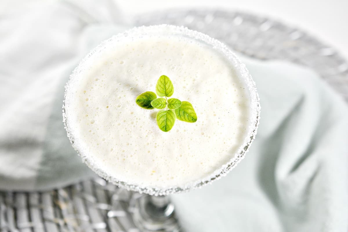 A frosty white Winter Keto Martini served in a keto sweetener rimmed martini glass, garnished with a small sprig of green basil leaves. The glass is set on a gray woven placemat with a light blue cloth in the background.
