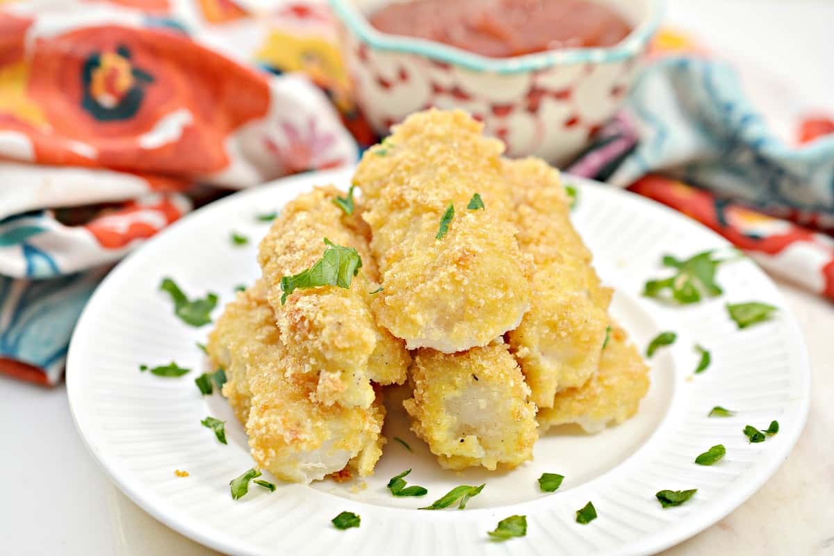 A white plate with as stack of golden-brown, breaded and baked keto fish sticks garnished with chopped parsley. In the background, there is a colorful napkin and a patterned bowl filled with ketchup.