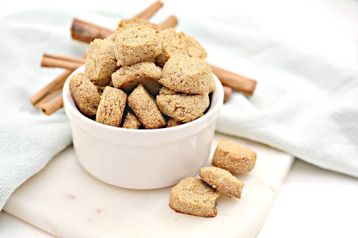 A white bowl filled with Keto Peppernuts is placed on a marble surface. A few cookies are scattered around the bowl, and several cinnamon sticks are arranged in the background, with a light blue cloth partially visible.