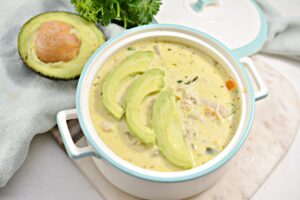 A white pot filled with Keto White Chicken Chili topped with sliced avocado. Behind the pot, there's a halved avocado and a bunch of fresh parsley on a white surface.