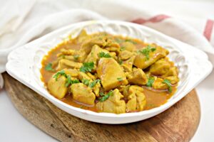A white ceramic plate filled with a vibrant, yellow-brown curried chicken dish, garnished with fresh cilantro. The plate sits on a round wooden cutting board. The background features a white cloth with a red stripe.