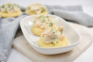 Two servings of Keto Chicken Salad Snacks are placed on top of crisp, rounds of keto fathead dough, displayed on a white plate. The chicken salad is mixed with bits of celery and mayonnaise, garnished with herbs. A gray and white patterned cloth is in the background.