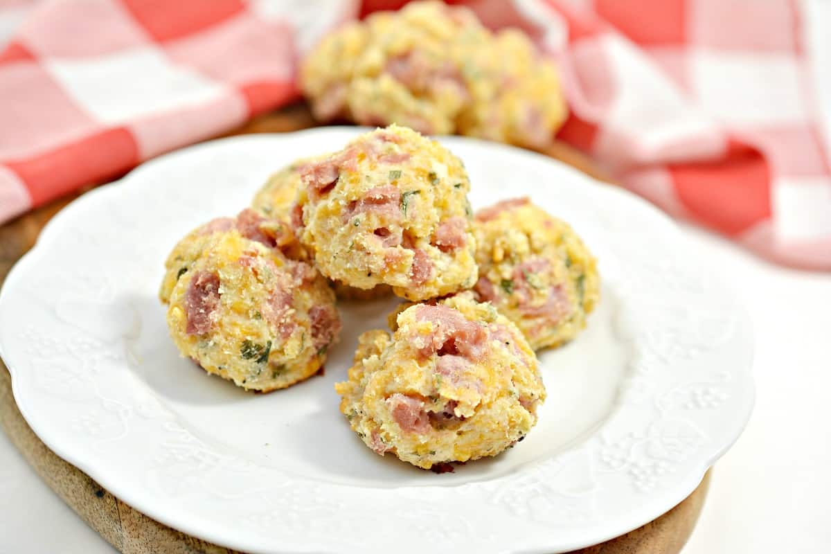 A white plate holds several keto ham and cheese bites placed on a wooden cutting board with a red and white checkered cloth in the background. These small, round appetizers feature visible pieces of ham and herbs mixed into the cheese.