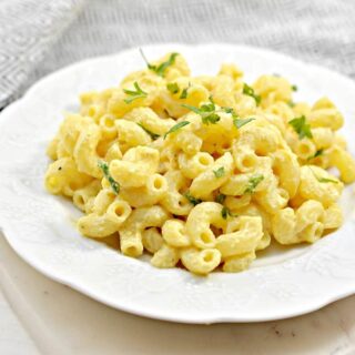 A white plate filled with creamy Cashew Mac and Cheese, garnished with parsley, sits on a white surface. A textured gray cloth is partially visible in the background.