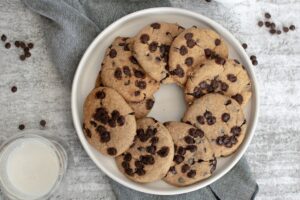 A white round plate filled with white bean chocolate chip cookies arranged in a circle. The plate is set on a grey cloth napkin, and there are scattered chocolate chips on a grey textured surface. A glass of milk is partially visible on the left side of the image.