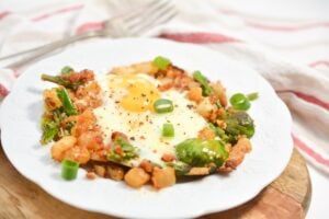 A white plate holds Low-Carb Keto Hash consisting of a sunny-side-up egg surrounded by diced potatoes, asparagus, Brussels sprouts, and chopped scallions. The meal is seasoned, and the plate is set on a wooden board with a fork and a red-striped towel in the background.