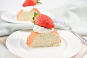 A slice of fluffy Keto Angel Food Cake topped with a dollop of whipped cream and a whole strawberry is placed on a white plate. In the background, there is another similar slice of cake and a fork resting on a napkin.