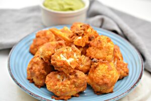 A blue plate filled with crispy, golden-brown buffalo cauliflower bites is placed on a table. A small white bowl of green dipping sauce is positioned in the upper left corner. A gray cloth napkin lies beside the plate.