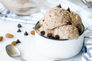 A bowl of creamy vegan chocolate ice cream topped with chocolate chips and chopped nuts, with a few scattered around. A spoon and a cloth napkin are beside the bowl. In the background, there is another bowl of the same ice cream.