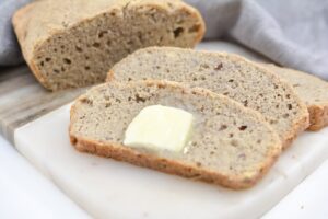A close-up of two slices of freshly baked Homemade Gluten Free Bread on a white cutting board. One slice has a pat of melting butter on top. A larger piece of the bread loaf is partially visible in the background, with a grey cloth nearby.