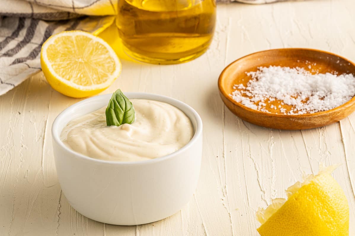 A white bowl filled with mayonnaise garnished with a basil leaf sits on a textured white surface. A halved lemon, a wooden bowl with salt, a bottle of olive oil, and a striped cloth are arranged around the bowl. A lemon peel is in the foreground.
