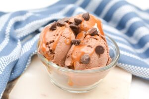 A glass bowl filled with three scoops of keto chocolate ice cream is garnished with keto chocolate chips and drizzled with caramel sauce. The bowl is placed on a white marble surface with a blue and white striped cloth in the background.