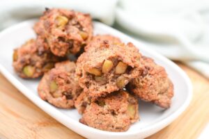 A white dish on a wooden surface holds several chunky, homemade Keto Apple Fritters. The fritters appear to be freshly baked, with a crumbly texture and visible pieces of apple embedded within them. A soft white cloth is in the background.