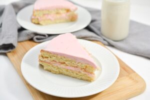 A slice of layered Keto Vanilla Cake with light pink frosting is elegantly displayed on a white plate. The layers of the cake are visible, showing a generous amount of frosting between them. Another slice is blurred in the background on a similar plate.