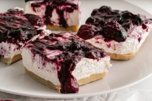 Four squares of No-Bake Keto Cheesecake sit on a white plate. The squares consist of a crumbly base, a thick layer of white cream cheese filling, and a generous topping of mixed berry compote, featuring dark purple-black berries.