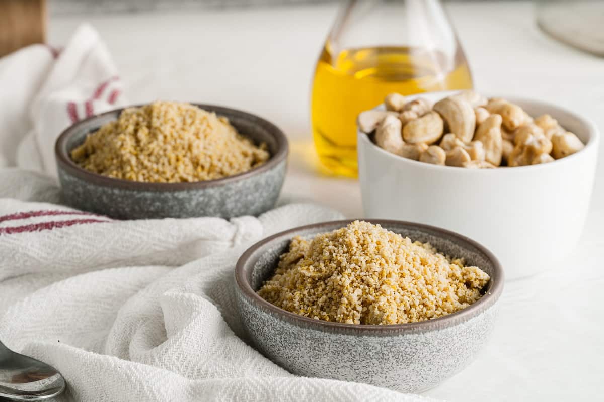 Two gray stone bowls with Vegan Parmesan Cheese are in focus in the foreground. Behind them, a white bowl filled with whole cashews and a glass bottle of oil are also visible. A white cloth with red stripes lies beside the bowls on a white surface.