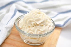 A glass bowl filled with piped Keto Vanilla Buttercream Frosting sits on a wooden board. The swirled peaks of the whipped cream are prominent and fluffy. A white and blue striped cloth is visible in the background.